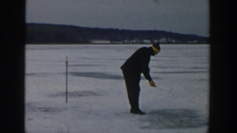 The man leans forward on the frozen Wisconsin lake, appearing to check or work at a spot in the ice with a small tool.