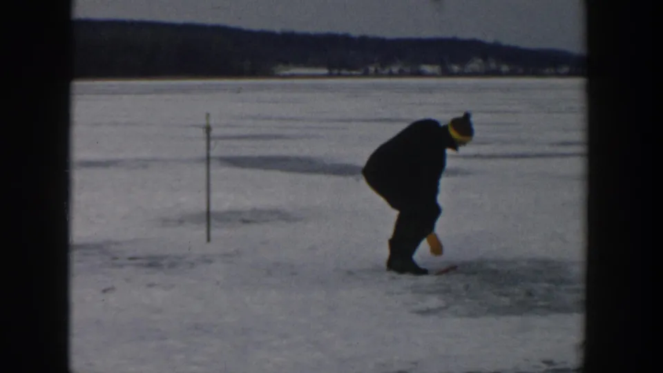 A man crouches over the frozen lake ice in Wisconsin, using or positioning a tool near a possible ice-fishing hole.