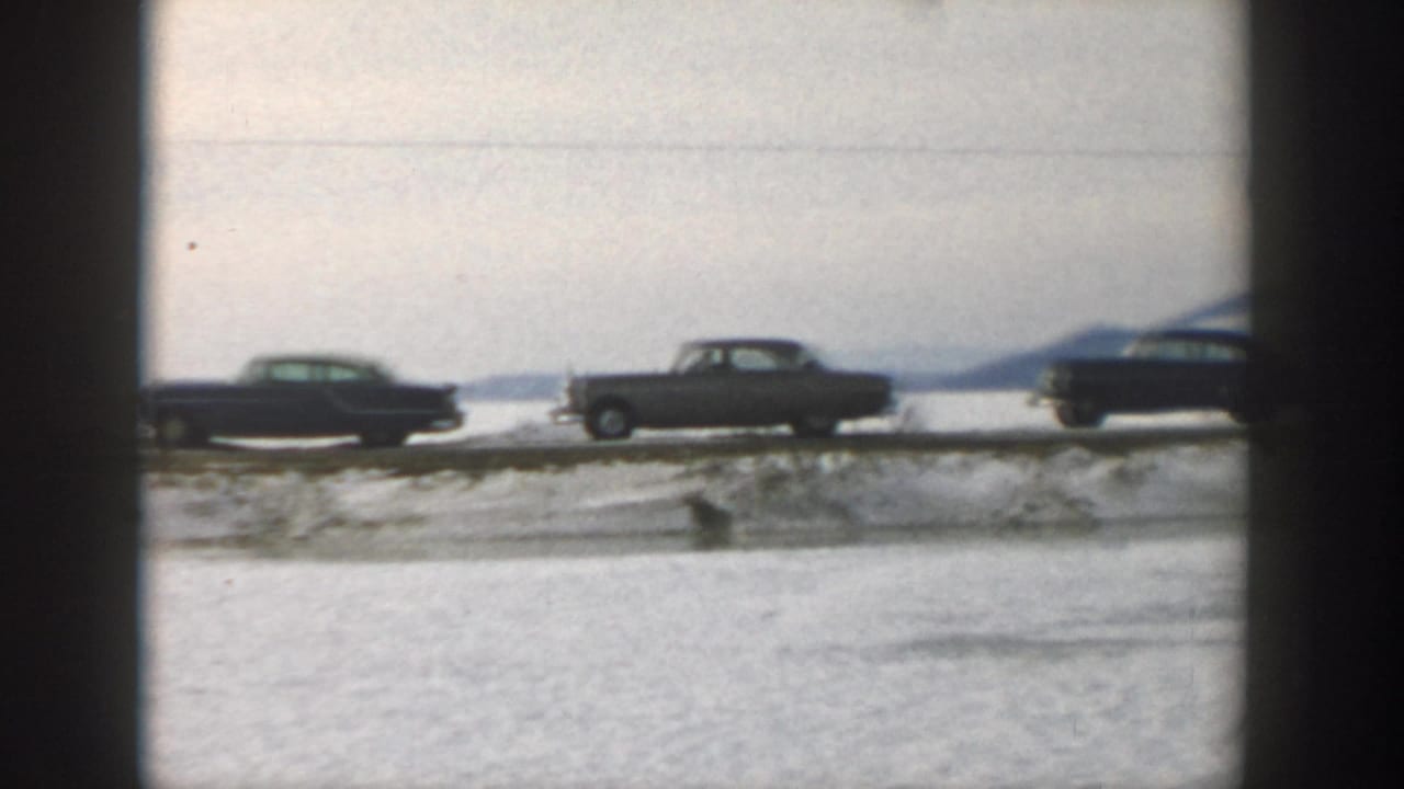Classic cars parked along a snowy roadside beside a frozen lake.