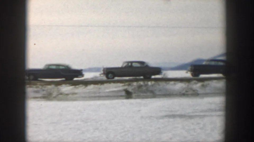 1957 Wisconsin winter home-movie frame with mid-century cars along a snowy road near a frozen body of water.