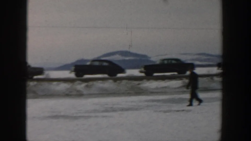 A distant figure walks across snow while 1950s cars sit along a Wisconsin roadside near a frozen lake.