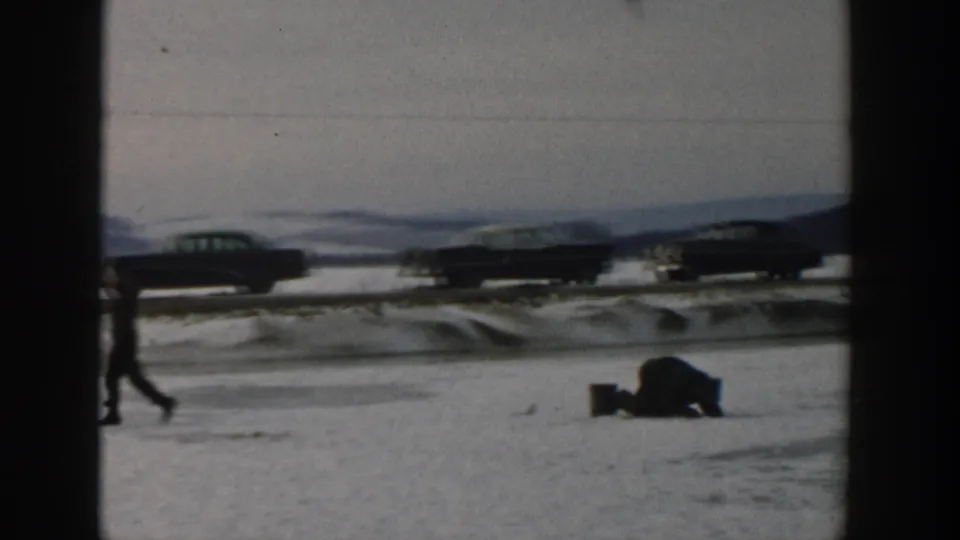 A winter home-movie frame shows a walker and a bundled figure low on the snow, with classic cars behind them in Wisconsin.
