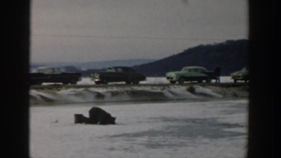 Mid-century cars, including a green car, stand along a snowy Wisconsin roadside near a frozen lake in 1957 footage.