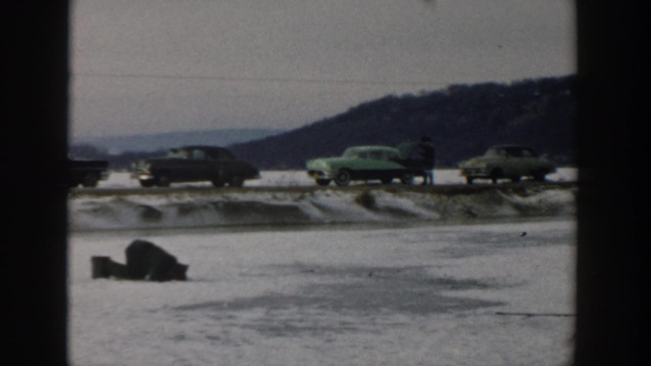 Frozen lake with a bundled person near the ice and classic cars passing on a snowy road.