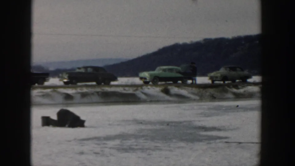 Wisconsin 1957 8mm frame showing a frozen lake, a bundled figure on the ice, classic cars, snow, and a wooded hill beyond the road.