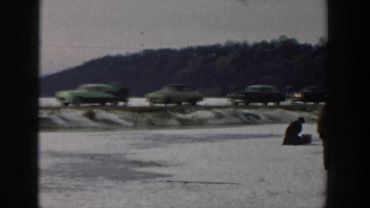 Wisconsin 1957 winter frame with a seated ice fisher on the frozen lake and several mid-century cars passing beside a wooded hill.