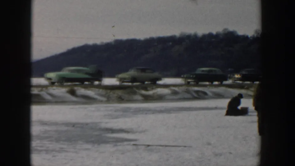 Wisconsin 1957 winter frame with a seated ice fisher on the frozen lake and several mid-century cars passing beside a wooded hill.