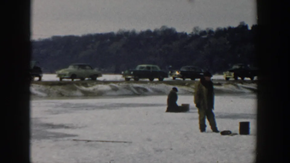 Wisconsin 1957 8mm frame showing two people ice fishing on a snowy frozen lake, with buckets, passing cars, and a wooded hill behind them.