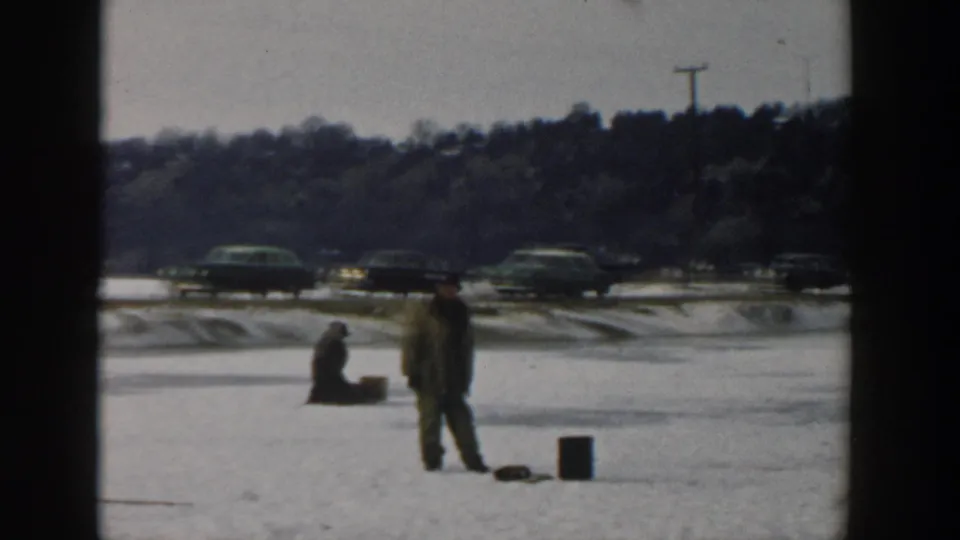 Wisconsin 1957 frame with one person standing and another seated on the frozen lake, ice-fishing gear nearby, and classic cars on the road behind.