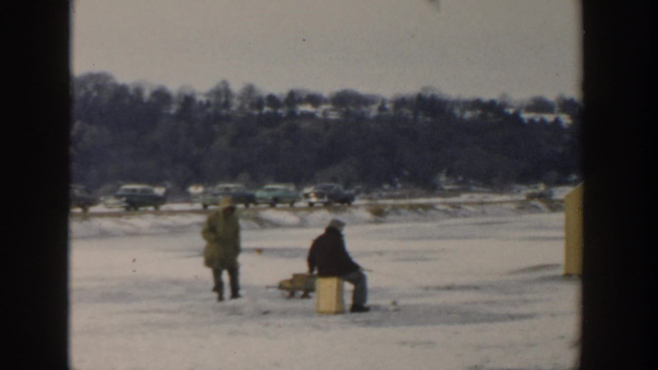 Two bundled people ice fish on a frozen lake with parked cars and snowy hills behind them.