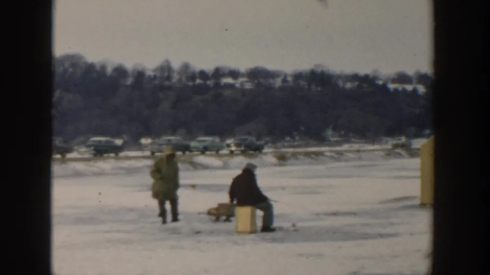 1957 Wisconsin home-movie frame showing two bundled anglers on a frozen lake near a bucket, with classic cars along the snowy shore.