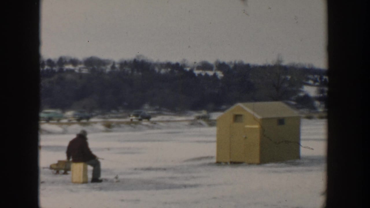 A seated ice fisher and yellow hut sit on a snow-covered frozen lake with cars in the distance.