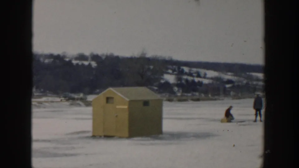 A yellow ice-fishing hut stands on a frozen Wisconsin lake in 1957, with two distant bundled figures working near the ice.