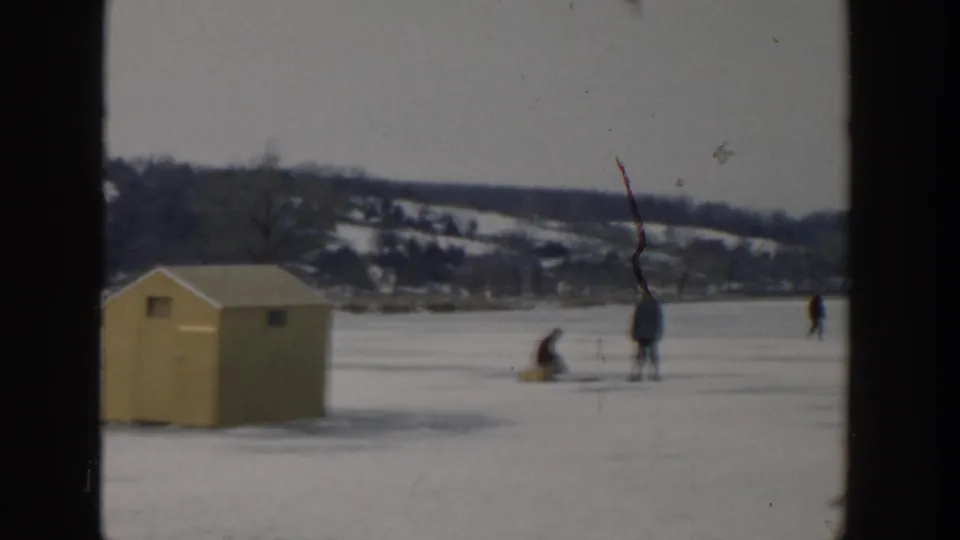 1957 Wisconsin winter frame showing a yellow ice hut, two distant anglers, and a broad frozen lake with snowy hills beyond.
