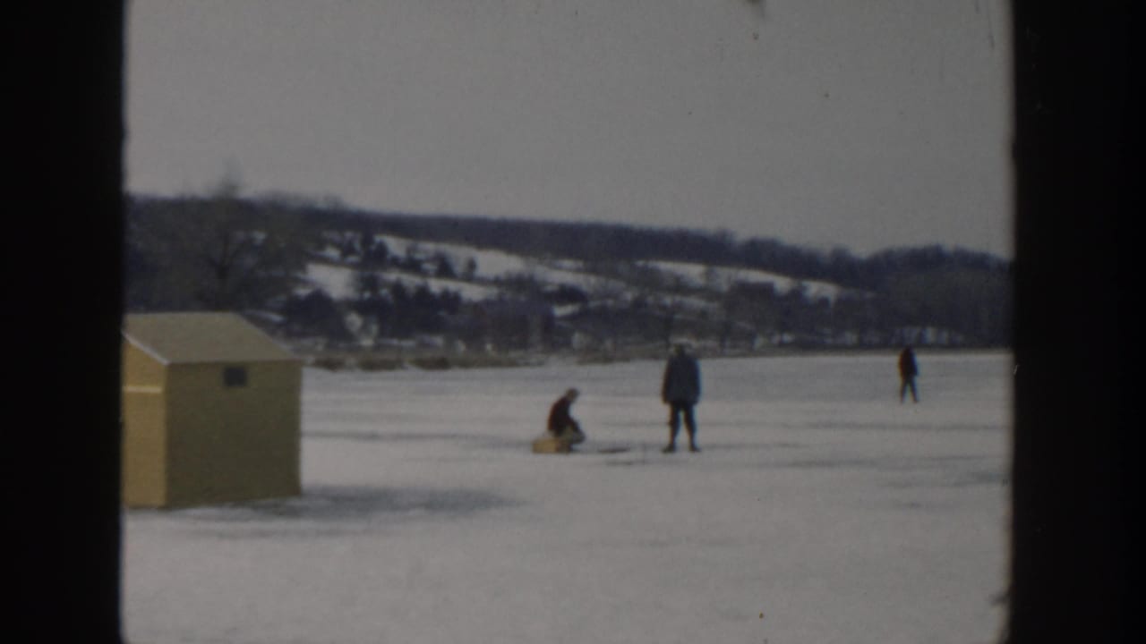 People ice fishing on a frozen lake near a yellow shed with snowy hills in the distance.