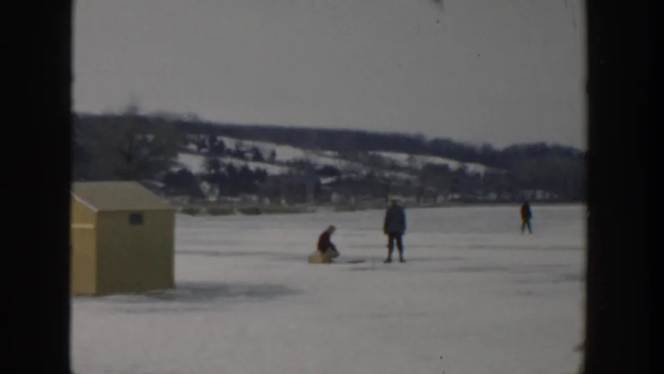 1957 Wisconsin home-movie frame showing bundled people ice fishing beside a yellow shed on a frozen lake.