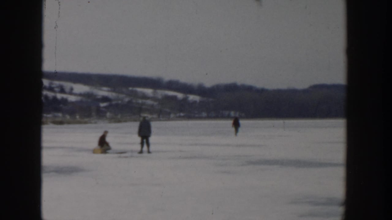 Bundled figures stand and crouch on a frozen lake with distant shoreline and hills.