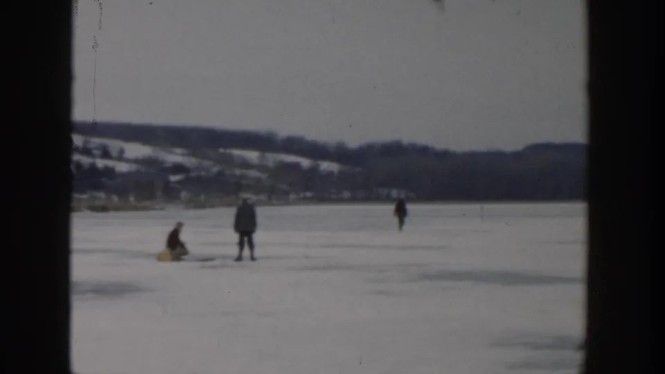 1957 Wisconsin frame of people spaced across a frozen lake, including one crouched near ice-fishing gear.