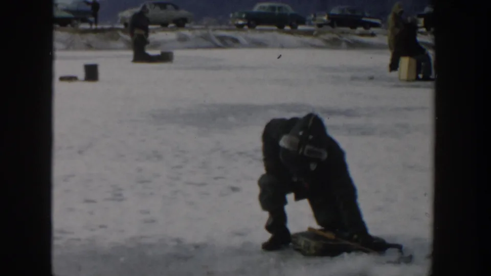 Closer 1957 Wisconsin frame showing a bundled angler crouched by gear on the ice, with parked cars behind.