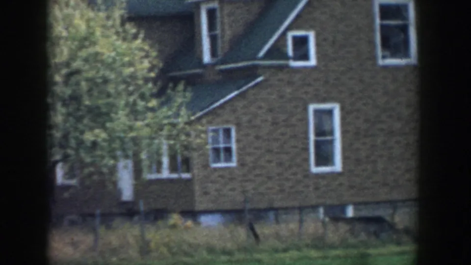 1958 Michigan home-movie frame of a brown brick house exterior with a white door, windows, trees, and backyard growth.