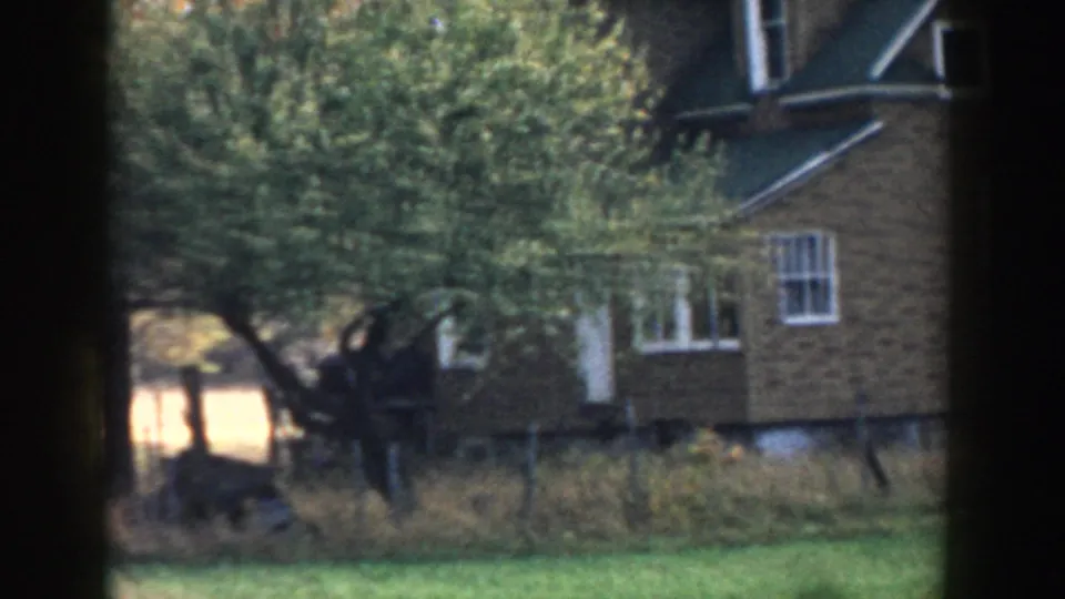 1958 Michigan 8mm frame showing the house partly obscured by trees, with grass, brush, and sunlit yard beyond.