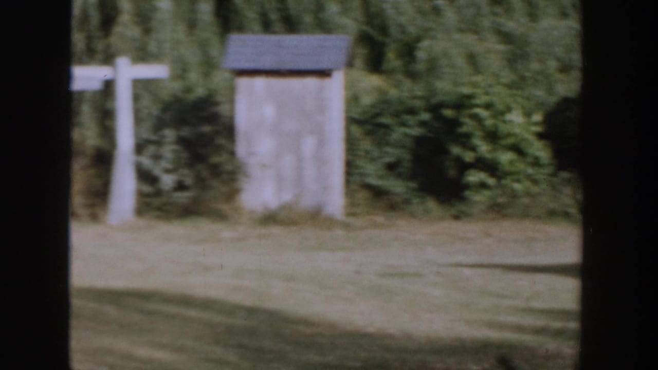 Small gray outbuilding near white fence posts and trees.