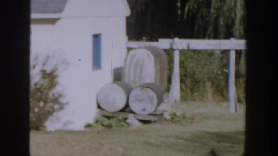 1958 Michigan 8mm frame showing barrels beside a white house wall, with a fence and trees beyond the yard.
