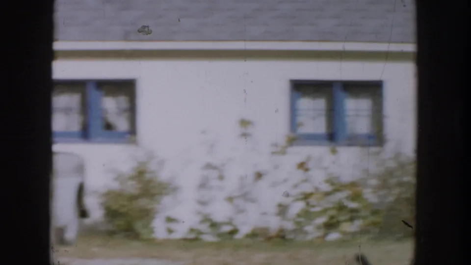 1958 Michigan home exterior with blue-trimmed windows, pale siding, shrubs, grass, and soft 8mm motion blur.