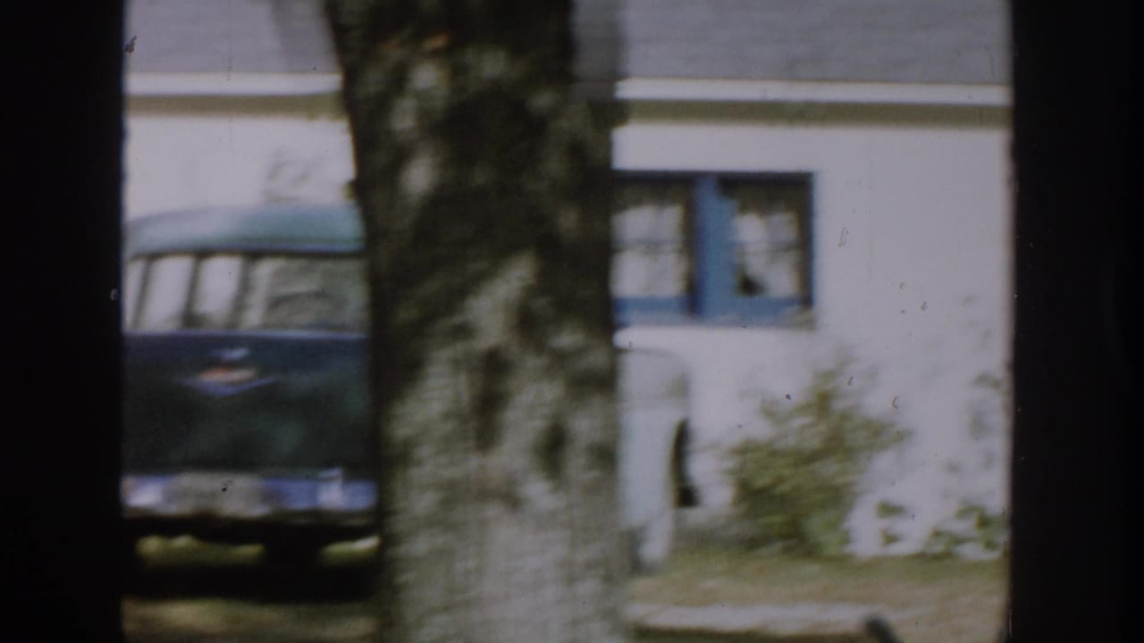 1958 Michigan home-movie frame showing a white house with blue trim, a tree trunk in front, and a dark mid-century car parked nearby.