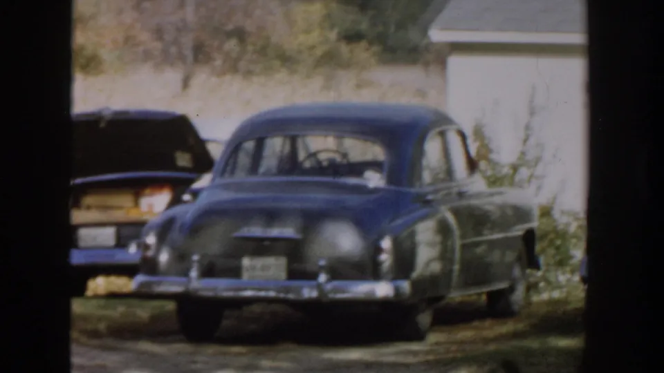 1958 Michigan frame with dark sedans and chrome trim parked beside a white house, seen through foreground trees.