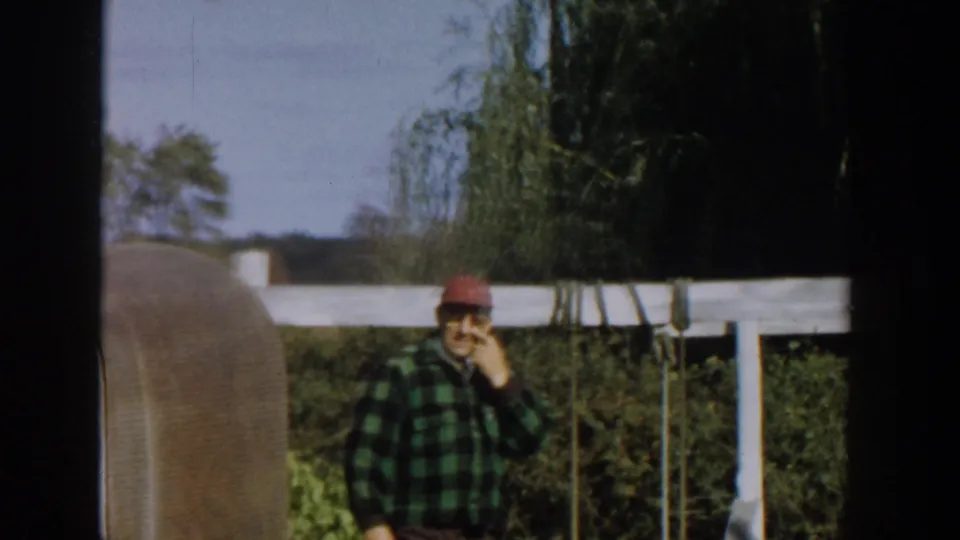 1958 Michigan frame showing an adult man in a green plaid shirt and red cap standing near a white fence, apparently smoking in a yard.