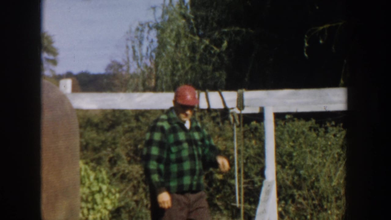 Man in a green plaid shirt and red cap walking beside a white fence in a leafy yard.