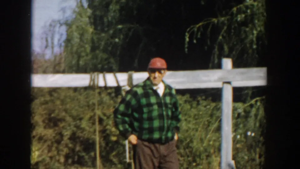 An adult man in a green plaid shirt, red cap and sunglasses stands or walks near a white fence in the Michigan yard setting.
