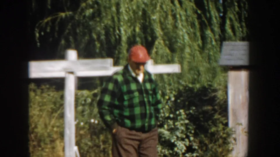 The man continues through the yard in a green plaid shirt, red cap and sunglasses, framed by a white fence and garden vegetation.