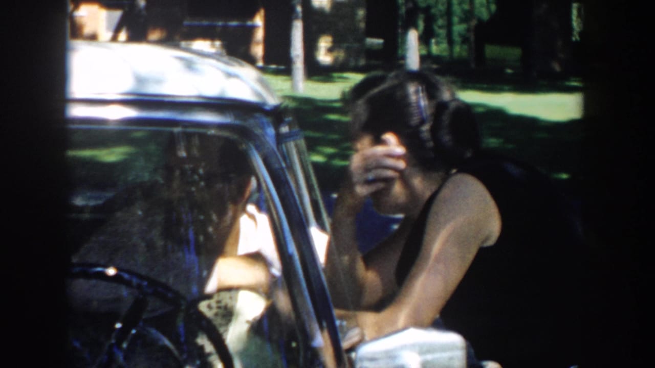 In 1958 Michigan home-movie footage, a woman leans into a parked car window while a man sits inside in bright outdoor light.
