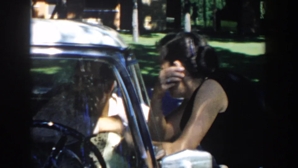 In 1958 Michigan home-movie footage, a woman leans into a parked car window while a man sits inside in bright outdoor light.