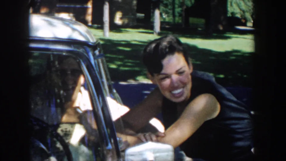 The woman smiles broadly beside the car window as she talks with the seated man in a sunlit 1958 Michigan home-movie scene.