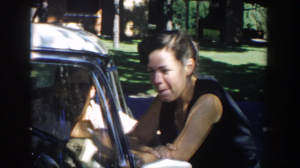 A woman in a dark sleeveless dress leans on the car window frame, continuing a close conversation in the outdoor Michigan setting.