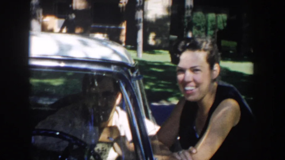 The woman glances toward the camera beside the car window, with the seated man still partly visible inside the 1958 Michigan scene.