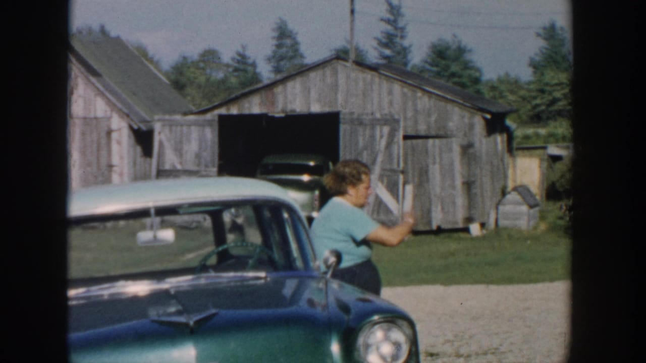 In 1958 Michigan home-movie footage, a woman in a teal shirt sits by a classic car with rural barns behind her.