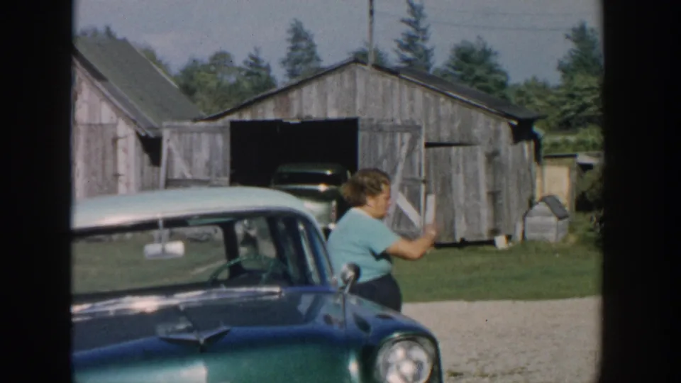 In 1958 Michigan home-movie footage, a woman in a teal shirt sits by a classic car with rural barns behind her.