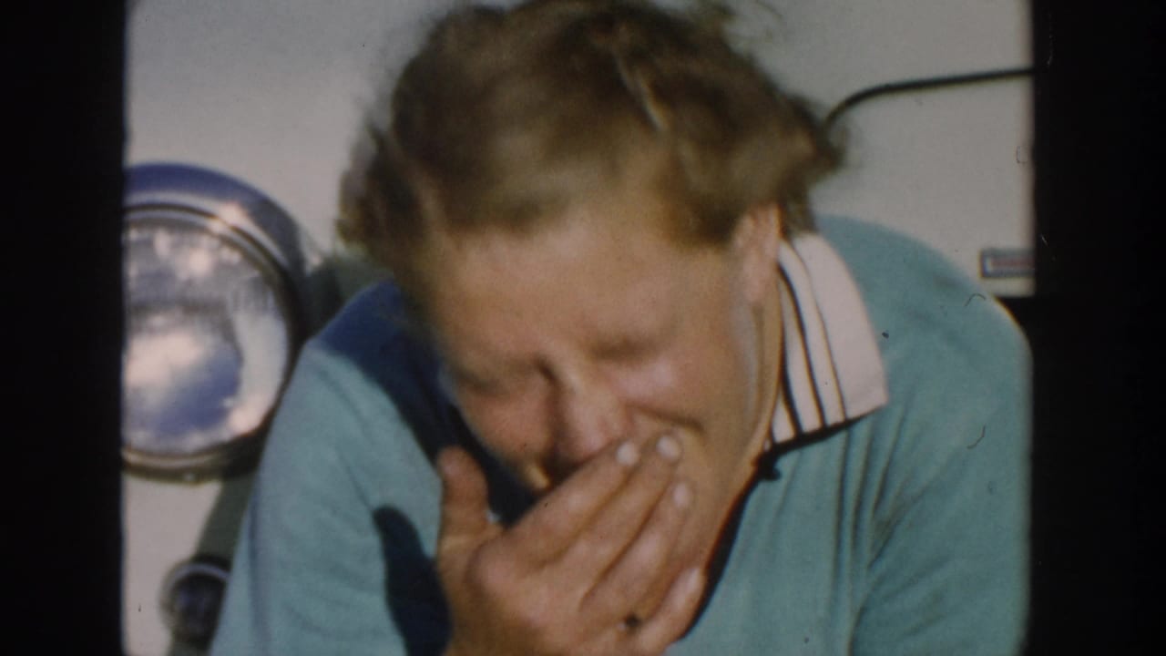 A blonde woman in a teal collared shirt covers her mouth while seated beside a classic car headlight in Michigan footage from 1958.
