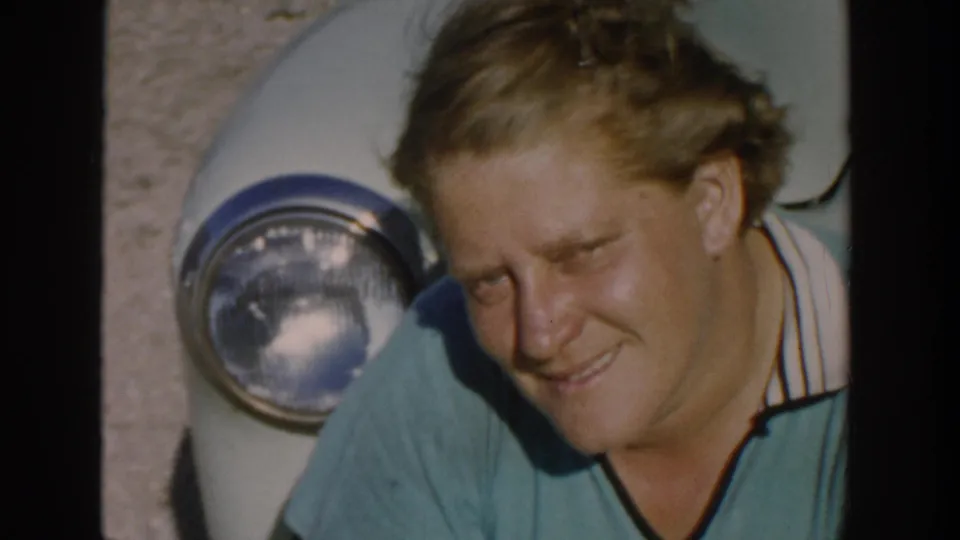 A woman smiles shyly beside a round car headlight, with grain and soft color visible in the 1958 Michigan home-movie frame.