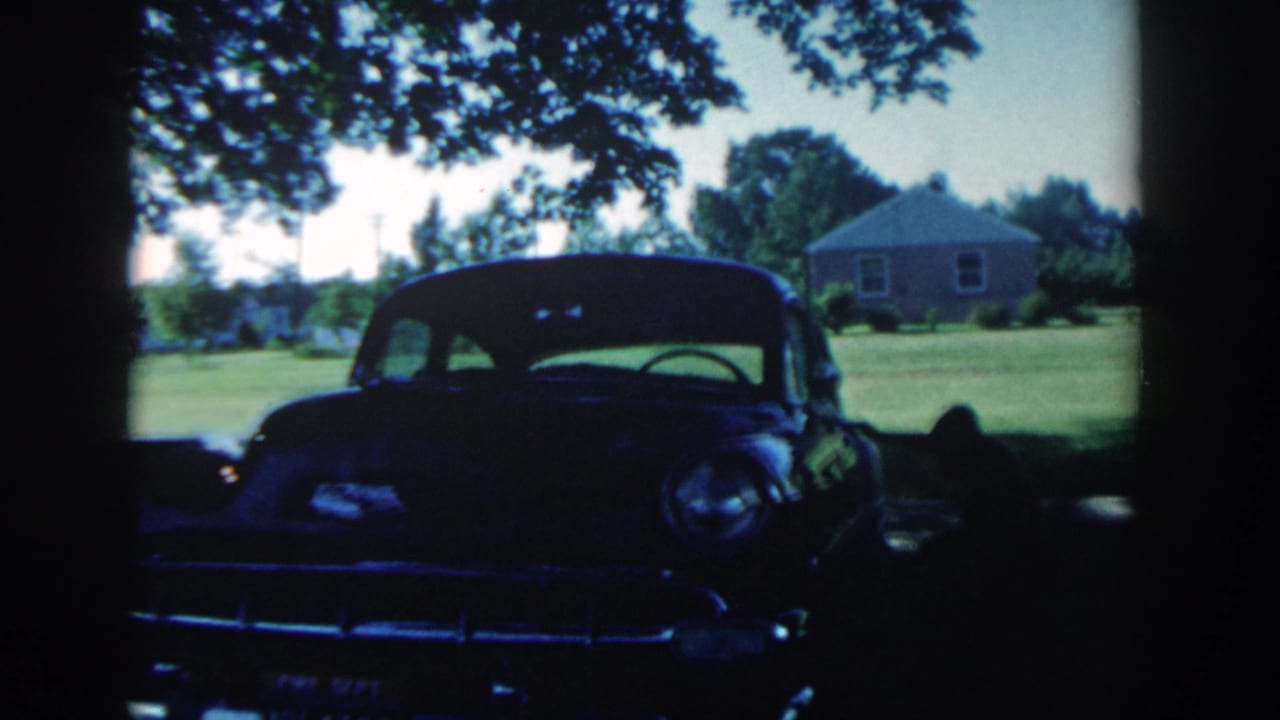 Front view of a dark classic sedan parked in shade with a lawn and house behind it.