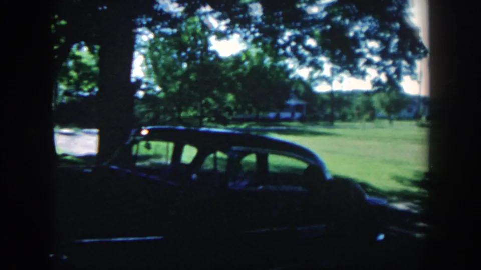 1958 Michigan home-movie view of a dark sedan parked near shade trees in a broad suburban yard.