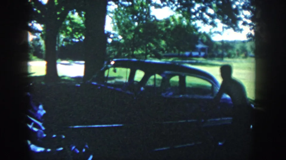 1958 Michigan frame showing a shirtless man beside the rear side of a dark classic sedan in a shaded yard.