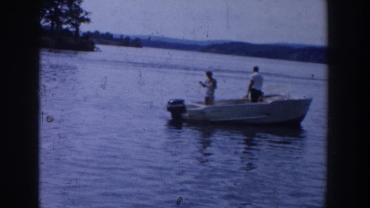 1958 Wisconsin 8mm frame showing two people standing in a small outboard motorboat on calm lake water.