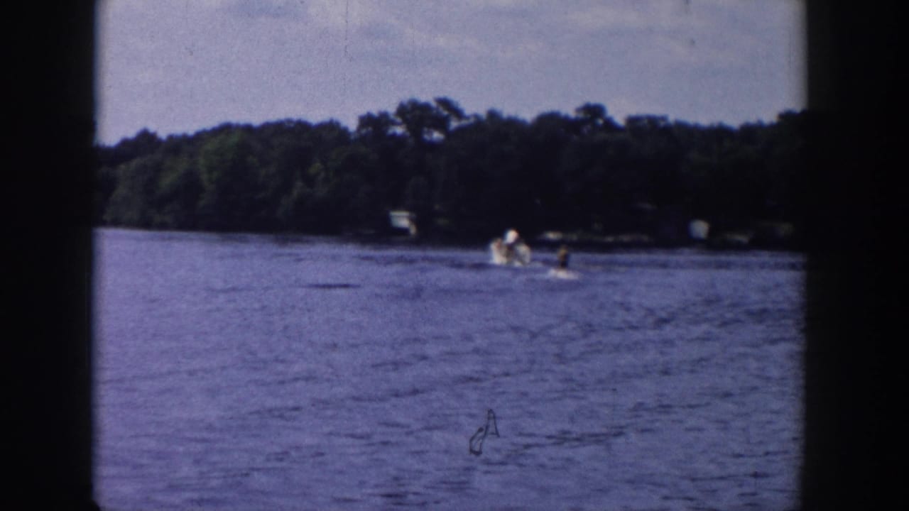 Water skier trails behind a boat on a lake near a wooded shoreline.