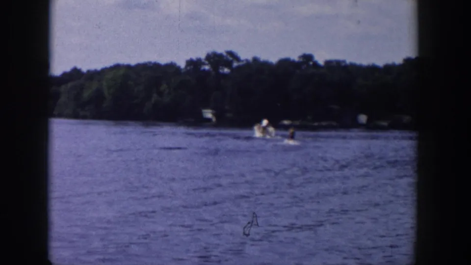 1958 Wisconsin 8mm frame showing a distant water skier being pulled across a lake near a tree-lined shore.