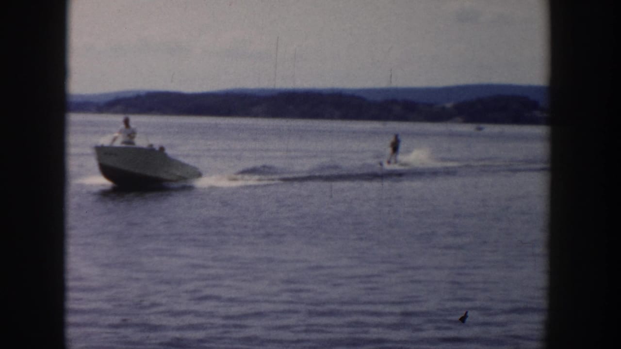 White motorboat pulls a water skier across a lake with distant wooded shoreline.